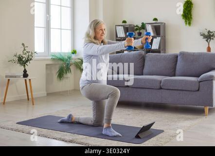 Donna anziana più anziana che si inginocchia, tiene i manubri, si allena, si allena, si guarda il tablet online Foto Stock