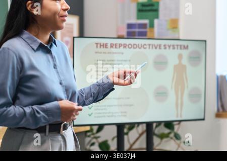Giovane donna ispanica adulta che presenta un diagramma del sistema riproduttivo su schermo digitale, tenendo un marcatore e gesti mentre spiega i contenuti educativi in aula Foto Stock