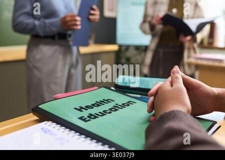 Adolescente seduto alla scrivania tenendosi per mano insieme con una cartella di educazione sanitaria in primo piano, due adulti in piedi in background tenendo documenti in aula Foto Stock