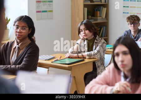 Gruppo di adolescenti multietnici seduti alle scrivanie ascoltando attentamente l'insegnante in classe, con quaderni e materiale scolastico sulle scrivanie, espressioni mirate che indicano il coinvolgimento nella lezione Foto Stock