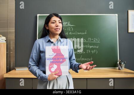 Donna ispanica in piedi in classe tenendo un libro di gravidanza, gestando mentre spiega le fasi di sviluppo prenatale davanti alla lavagna con note della lezione Foto Stock