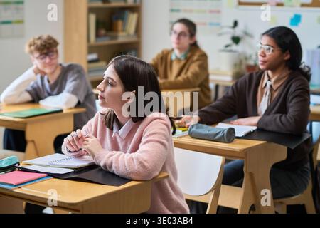 Gruppo di adolescenti multietnici seduti alle scrivanie ascoltando attentamente durante la lezione in aula, concentrandosi sull'insegnante fuori dalla cornice, sui quaderni e sulle forniture scolastiche sulle scrivanie, imparando in ambiente educativo Foto Stock