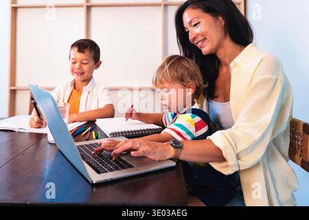 Famiglia asiatica a casa - madre asiatica che lavora in multitasking a casa, lavora su un notebook e aiuta i suoi figli a disegnare, fare i compiti e imparare al tabl Foto Stock