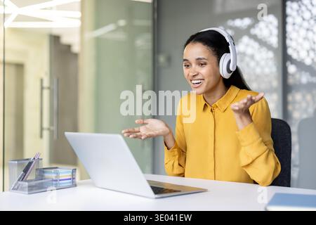Giovane donna sorridente che sta facendo una divertente videochiamata online, comunica attivamente con i gesti delle mani indossando le cuffie bianche e seduto alla scrivania dell'ufficio con un laptop Foto Stock
