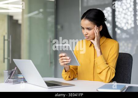 Giovane donna professionale che si sfrega da un mal di testa mentre esamina un tablet su una scrivania con un notebook, mostrando stress, esaurimento e pressione lavorativa in un moderno ambiente di ufficio Foto Stock