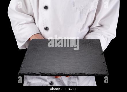 Lo chef in uniforme bianca regge un piatto da portata vuoto in ardesia nera. Sfondo nero. Foto Stock