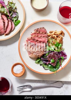 La tenera bistecca di manzo cotta viene tagliata a fette e posta su un piatto accanto a un'insalata verde e ai funghi. Due bicchieri di vino rosso seduto nelle vicinanze. La funzione di impostazione Foto Stock