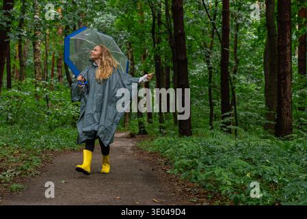 Una donna con stivali gialli e un impermeabile cammina con gioia su un sentiero nella foresta, circondato da una vegetazione lussureggiante, tenendo in mano un ombrello trasparente, magari godendosi Foto Stock