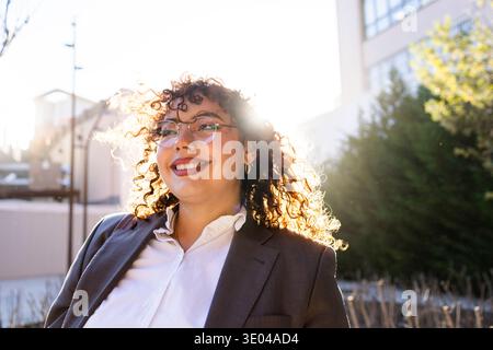Una donna d'affari sicura con capelli ricci e travi di occhiali con un sorriso luminoso. È vestita con una tuta formale e si trova in un ambiente urbano all'aperto Foto Stock