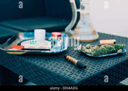 Un primo piano di un tavolo in vimini con boccioli di cannabis su un vassoio, un giunto arrotolato e vari accessori per fumatori, messi a confronto con posti a sedere informali all'aperto Foto Stock