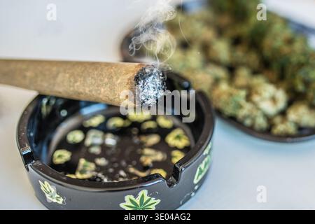 Primo piano di un giunto di cannabis illuminato che emette fumo accanto a un posacenere decorativo con disegni a foglia di cannabis, con gemme di cannabis sfocate sullo sfondo. Foto Stock