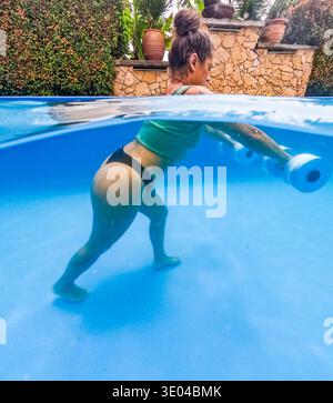 Donna impegnata nel fitness subacqueo usando pesi in acqua in una piscina, catturata in un ambiente vivace e chiaro. L'attività sottolinea la forza A. Foto Stock