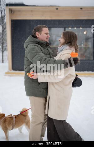 Una coppia amorevole si abbraccia in un paesaggio innevato, godendosi una tranquilla passeggiata invernale. Vestiti calorosamente, camminano con il loro piccolo cane dietro di loro, assaporando il Foto Stock