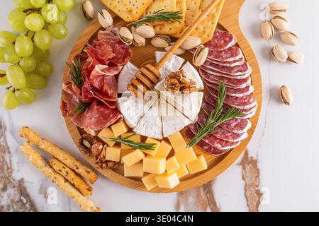 Un delizioso vassoio di formaggi con un assortimento di formaggi, affettati, cracker e uva fresca, guarniti con noci ed erbe aromatiche, ideale per il gour Foto Stock
