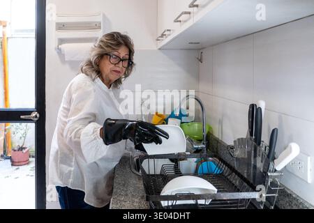 Donna anziana che lava i piatti in una cucina domestica, indossando guanti protettivi in gomma per lavori domestici Foto Stock