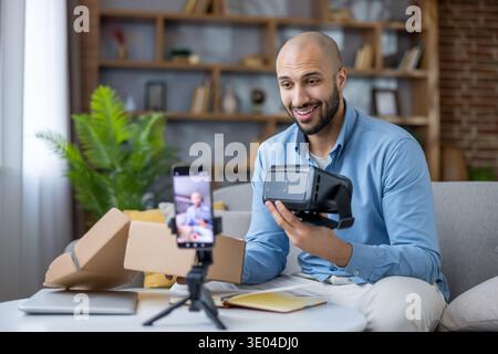 Uomo che registra un video per i social media sulla sua esperienza di unboxing e la recensione di un visore per la realtà virtuale, condividendo le sue opinioni sul nuovo Technolo Foto Stock