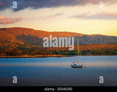 Barca a vela ancorata sulle acque calme della baia di Kenmare durante la luce dorata della sera nella contea di Kerry, Irlanda. Foto Stock