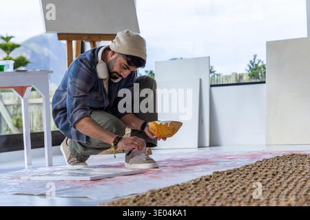 Uomo barbuto accovacciato su un foglio in studio, indossando un berretto, applicando vernice dalla ciotola alla tela del pavimento Foto Stock