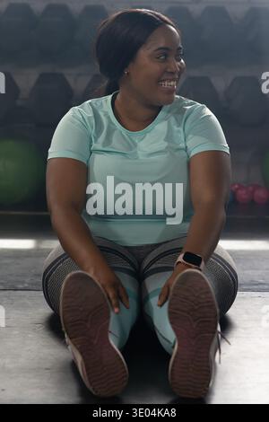Donna afroamericana seduta sul pavimento della palestra con gambe allungate, indossa camicia color verde acqua, smartwatch visibile Foto Stock