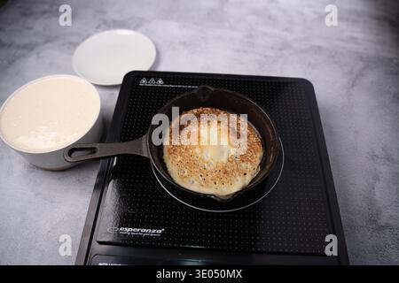 frittura di pancake marrone dorato in una padella di ghisa stagionata su un piano cottura a induzione nero, con una ciotola di pastella e un piatto bianco su un piatto di co testurizzato grigio Foto Stock