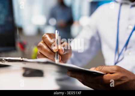 Primo piano del lavoratore che scrive sugli appunti mentre è seduto alla scrivania, svolgendo attività di contabilità. Dipendente nel moderno ambiente di lavoro che completa la documentazione durante le attività amministrative di routine Foto Stock