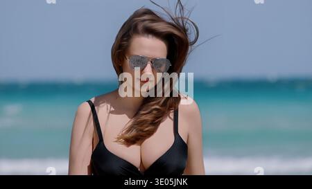 Vacanze estive e vacanze al mare Una giovane donna con costume da bagno nero e occhiali da sole aviatore gode di una vacanza tropicale in una giornata di sole con vista sull'oceano Foto Stock