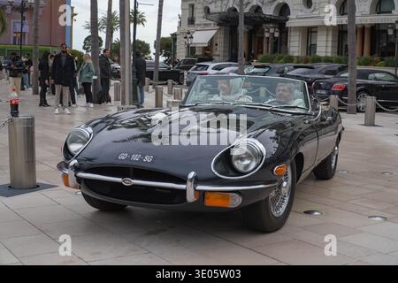 Classica Jaguar e-Type convertibile all'esterno dell'hotel le Louis XV, Place du Casino, Monaco, Francia. Una coppia più vecchia che guida un'auto di lusso attraverso la piazza trafficata. Foto Stock