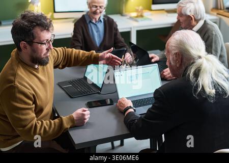 Giovane uomo adulto caucasico che insegna competenze informatiche di uomini e donne caucasici durante il corso di alfabetizzazione, gruppo seduto a tavola utilizzando computer portatili, apprendimento della tecnologia digitale insieme Foto Stock