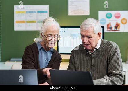 Donna caucasica anziana e uomo caucasico anziano che partecipano a un corso di alfabetizzazione informatica, seduti insieme alla scrivania, utilizzando computer portatili, donna che punta allo schermo, uomo che guarda attentamente Foto Stock