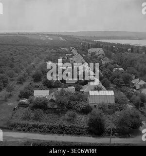 Un'impressionante fotografia vintage in bianco e nero degli anni '1980 presenta una vista panoramica sopraelevata del villaggio residenziale presso l'impresa Donrybkombinat nel quartiere di Slavyansk. L'ampia prospettiva aerea cattura file ordinate di tradizionali case rurali a un piano con giardini privati, circondati da lussureggianti alberi verdi, che si estendono verso un vasto e tranquillo bacino idrico. Questo paesaggio altamente autentico conserva splendidamente la serena vita quotidiana e l'atmosfera nel tranquillo Donbass prima della guerra. Foto Stock