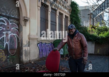 Un uomo con una giacca di pelle si erge su una strada della città con una sedia da ufficio rossa recuperata sullo sfondo di edifici industriali ricoperti di graffiti in una Foto Stock