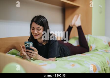 Giovane donna che si rilassa sul letto sorridente mentre usa lo smartphone e chatta a casa Foto Stock