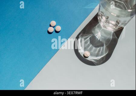 Un singolo tablet e un bicchiere di acqua fresca su uno sfondo minimalista blu bicolore. Concetto di dosaggio medico, recupero dei pazienti e benessere sanitario Foto Stock