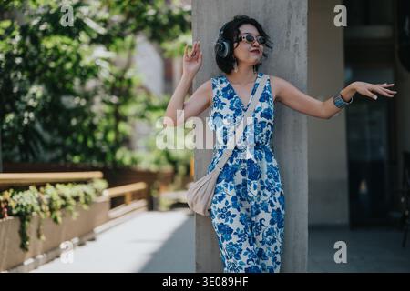 Donna in tuta floreale blu si appoggia su una colonna all'aperto, ascoltando le cuffie con stile Foto Stock