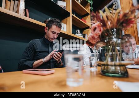 L'uomo in camicia nera usa lo smartphone in un bar con colleghi e decori con motivi floreali Foto Stock