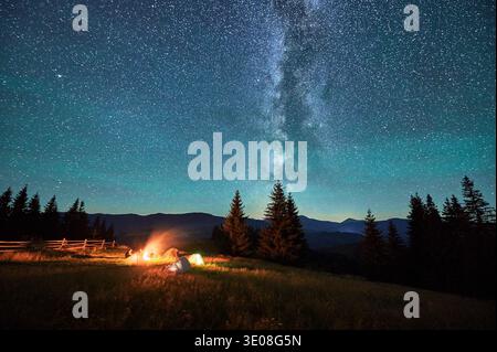 Sagome sfocate di turisti che riposano in campeggio intorno al fuoco con tende luccicanti nelle vicinanze sotto il cielo stellato. La via Lattea si estende in alto, gettando una luce magica sul tranquillo paesaggio montano. Foto Stock