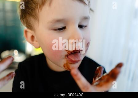 Un bambino di tre anni con cioccolato fuso sul viso. Vista ravvicinata che mette in risalto pelle ed espressioni disordinate. Concetto di trattamento vietato, dieta malsana, allergie, rischio di diabete. Foto Stock