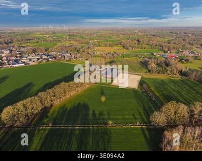 Vista aerea alta di un paesaggio olandese caratterizzato da scuderie di cavalli, pascoli verdi e una città lontana con turbine eoliche. Panoramica campagna rurale nel Foto Stock