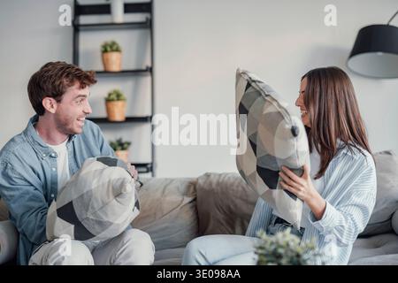 Allegra e varia coppia sposata innamorata seduto sul divano nel soggiorno di casa. Millennial abbastanza misto razza moglie e marito caucasico si divertono a giocare Foto Stock