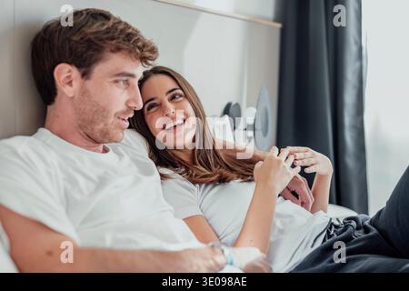 Una giovane coppia giace a letto, abbracciando e parlando in silenzio. I loro sorrisi e i loro tocchi delicati mostrano conforto, vicinanza e amore, condividendo una momen pacifica Foto Stock