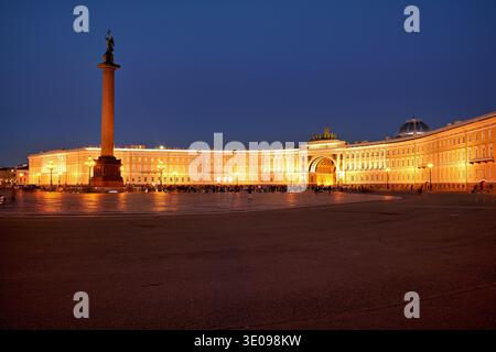 Il Palazzo d'Inverno si erge alto nella Piazza del Palazzo, illuminato con calore sotto il cielo notturno, con la colonna di Alessandro nelle vicinanze e la gente che cammina intorno. Foto Stock