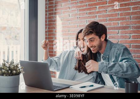 Una coppia millennial felicissima sedersi alla scrivania guardare lo schermo del notebook sentirsi euforici con la vittoria online, felici giovani marito e moglie entusiasti leggono buone notizie, Get Foto Stock
