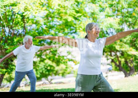 Attiva coppia senior che pratica yoga insieme in un parco verde lussureggiante, godendo di benessere, equilibrio e fitness all'aperto. Uomini e donne anziani che si esibiscono Foto Stock