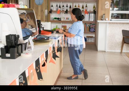 Una donna sta al bancone in un bar a preparare il cibo. Si concentra sul suo lavoro con decorazioni e altri oggetti visibili sullo sfondo. Foto Stock