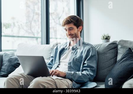 L'uomo bello e serio si siede sul divano con il computer in giro, passa il tempo a navigare sul Web a casa, usa la tecnologia moderna, fai ordine su Internet. Freelanc Foto Stock