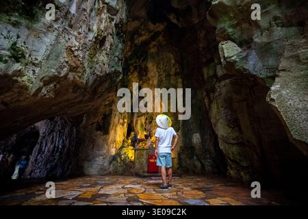La persona che indossa un tradizionale cappello conico vietnamita si erge dietro la macchina fotografica, osservando una statua e un altare luminosi all'interno di un vasto e antico natur Foto Stock