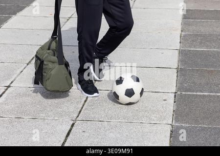 Persona in piedi su piastrelle plaza con pantaloni neri e sneakers nere con palla da calcio e borsone Foto Stock