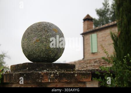 Ornamento a sfera in pietra intemprata su pareti in mattoni rustici con tradizionale casa di campagna e persiane verdi sullo sfondo nella campagna Toscana, Italia. Foto Stock