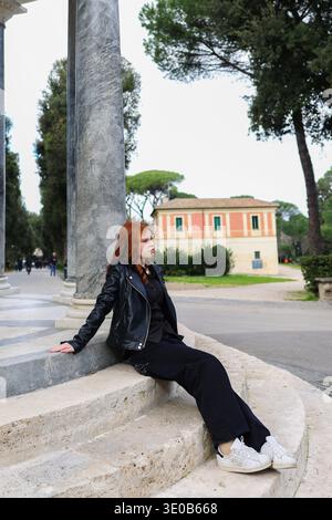 Giovane donna dai capelli rossi seduta accanto a colonne di marmo nello storico parco europeo. Viaggia con un'architettura classica. Foto Stock