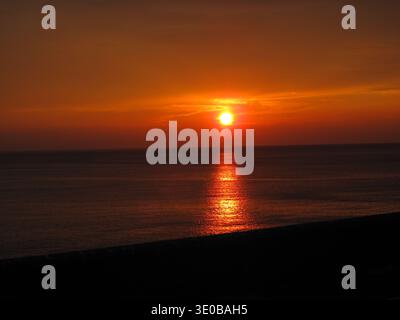 Un radioso tramonto sul mare, con toni arancioni e rossi che si riflettono sull'acqua calma. Il sole siede basso all'orizzonte. Foto Stock